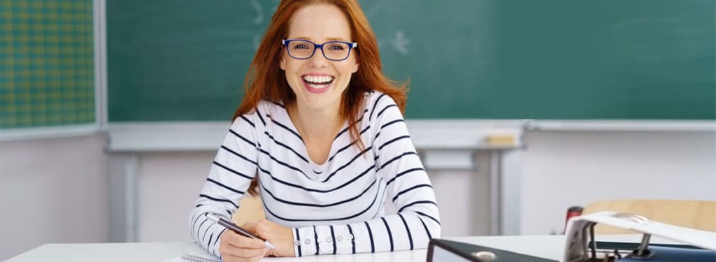 Lernende Frau mit Brille im Klassenzimmer, lächelnd und bereit für den Unterricht.