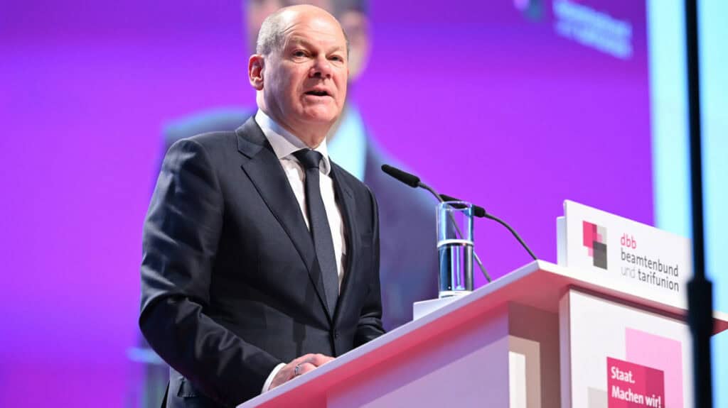 Olaf Scholz bei einer Rede auf einer Konferenz, im Hintergrund ein Bildschirm mit Logo.