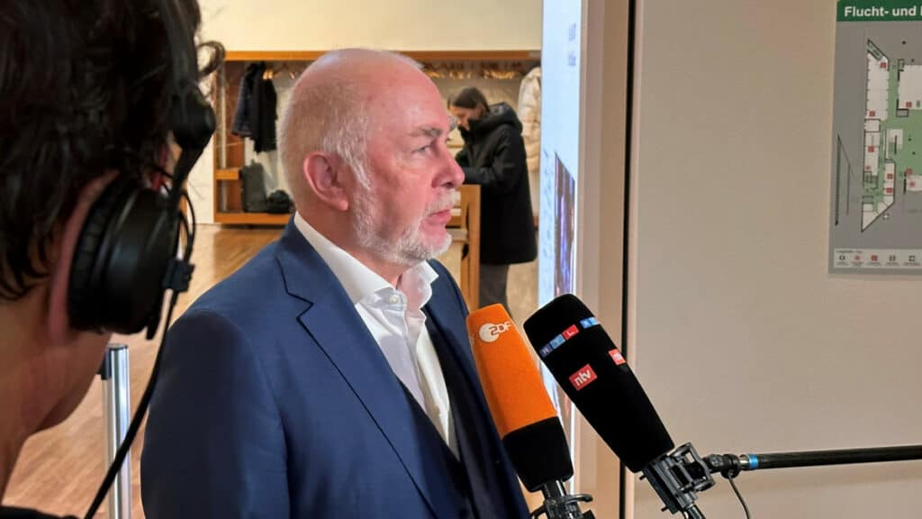 Politiker im Interview zu Bezahlungsrückständen in Ländern, mit Mikrofonen und Medien im Hintergrund.