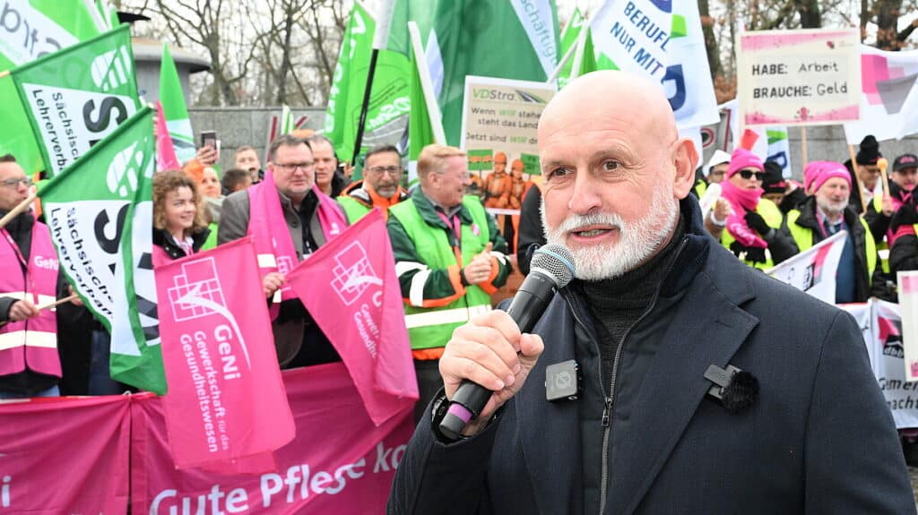 Mann spricht bei Protestdemo mit Mikrofon, Demonstranten mit Fahnen im Hintergrund.